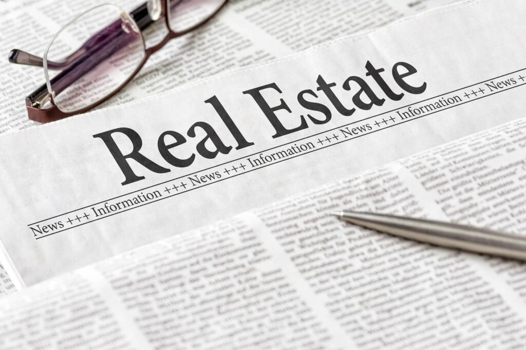 A pair of glasses and a pen resting on a newspaper section titled "real estate".